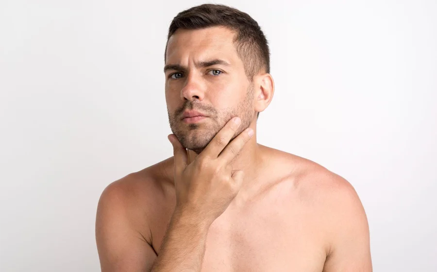 Confident shirtless man with a well-defined jawline examining his face, representing the results of male jawline contouring treatment